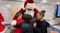  Two male and one female student hugging Santa in front of a classroom whiteboard.